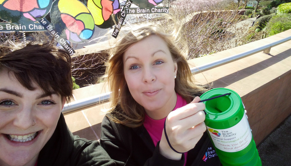 Two young women in Brain Charity outfits hold up a collection tin