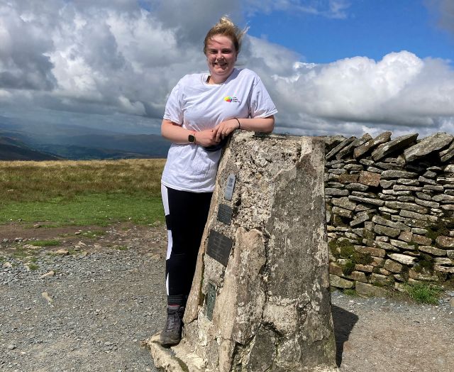 Woman standing at a peak marker in sunshine during the Yorkshire Three Peaks challenge, wearing a Brain Charity t-shirt.