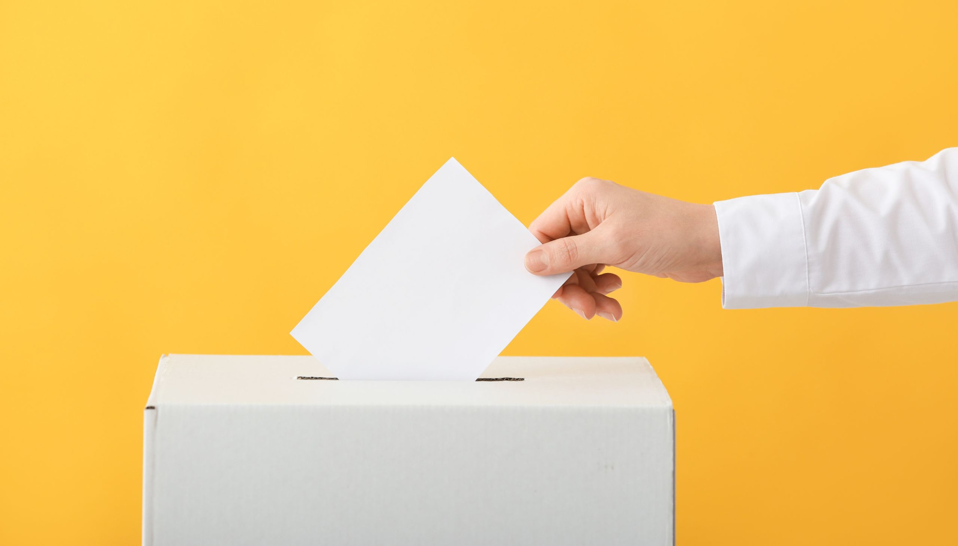 Hand placing a voting slip into a ballot box against a yellow background
