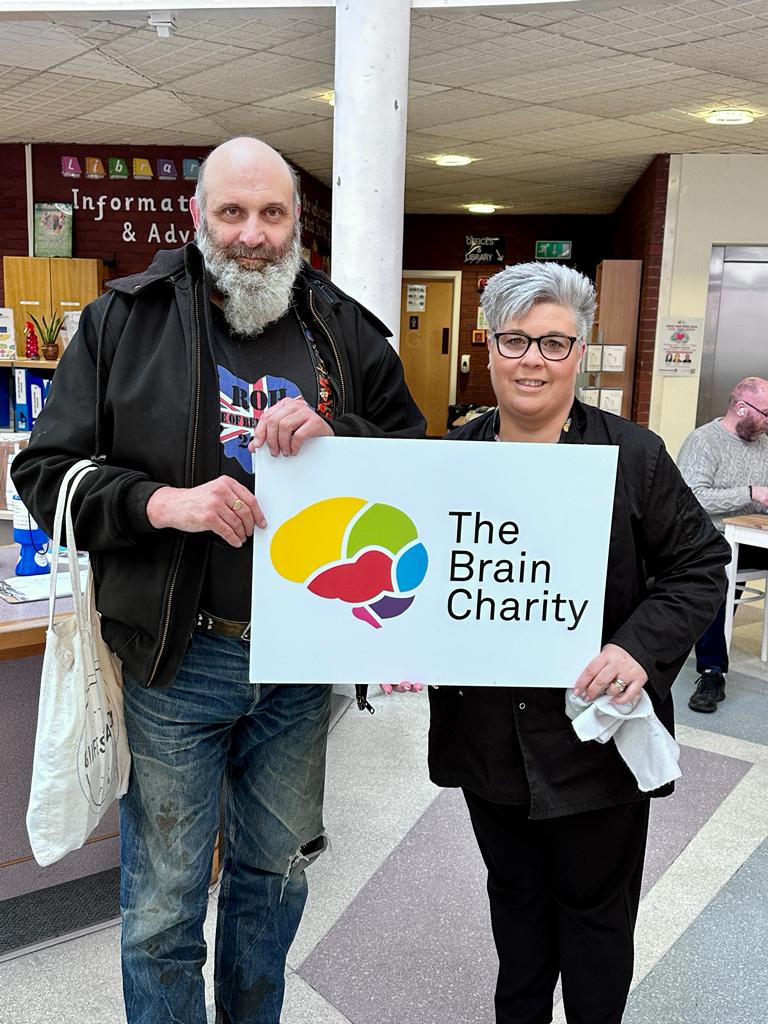 Fundraiser Ian from Bikers for Neuro visiting The BRain Charity to deliver his donation. He is standing iat our reception with a staff member and they ate holing a large sign with The Brain Charity logo on it