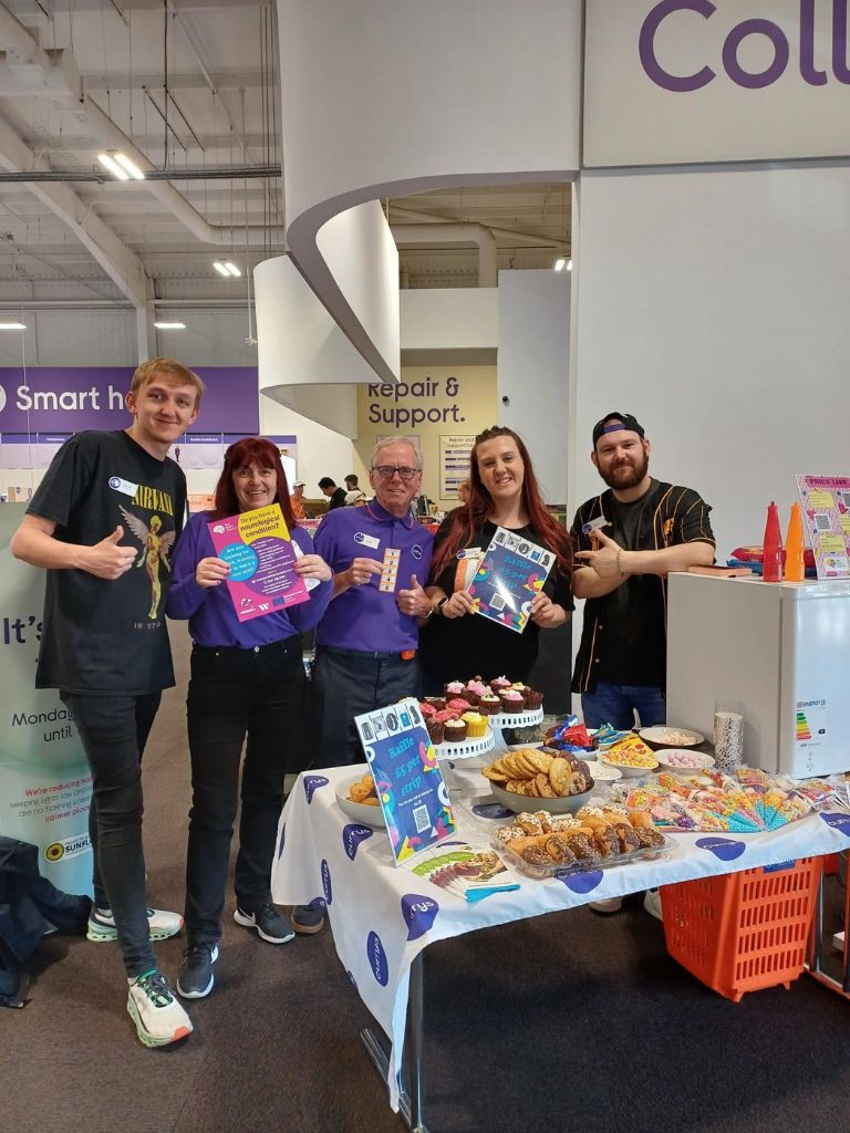 Amanda and her team wearing 90s gear at their workplace, Currys Aintree. There is a table in front of them laden with delicious looking homemade cakes and biscuits along with awareness raising materials about The Brain Charity 