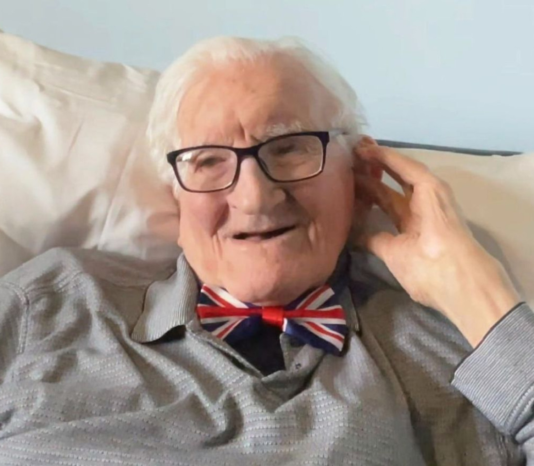 Photo of John wearing a grey shirt and a Union Jack bow tie. He's seated and holding his hand to his ear to be able to be able to hear better.