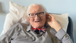 Photo of John wearing a grey shirt and a Union Jack bow tie. He's seated and holding his hand to his ear to be able to be able to hear better.