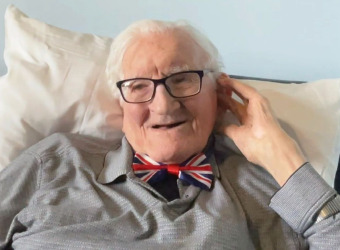 Photo of John wearing a grey shirt and a Union Jack bow tie. He's seated and holding his hand to his ear to be able to be able to hear better.