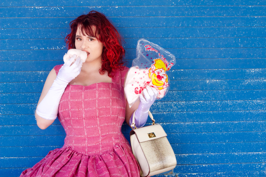Moray in a pink dress, standing in front of a blue wall. The dress has a square pattern made up from the words Moray Luke and eating candyfloss. She is holding a Moray Luke handbag.