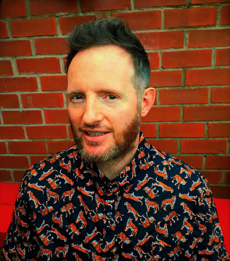 A bearded man with dark hair in a black shirt with small, colourful images of tigers on it. There is a plain brick wall in the background.