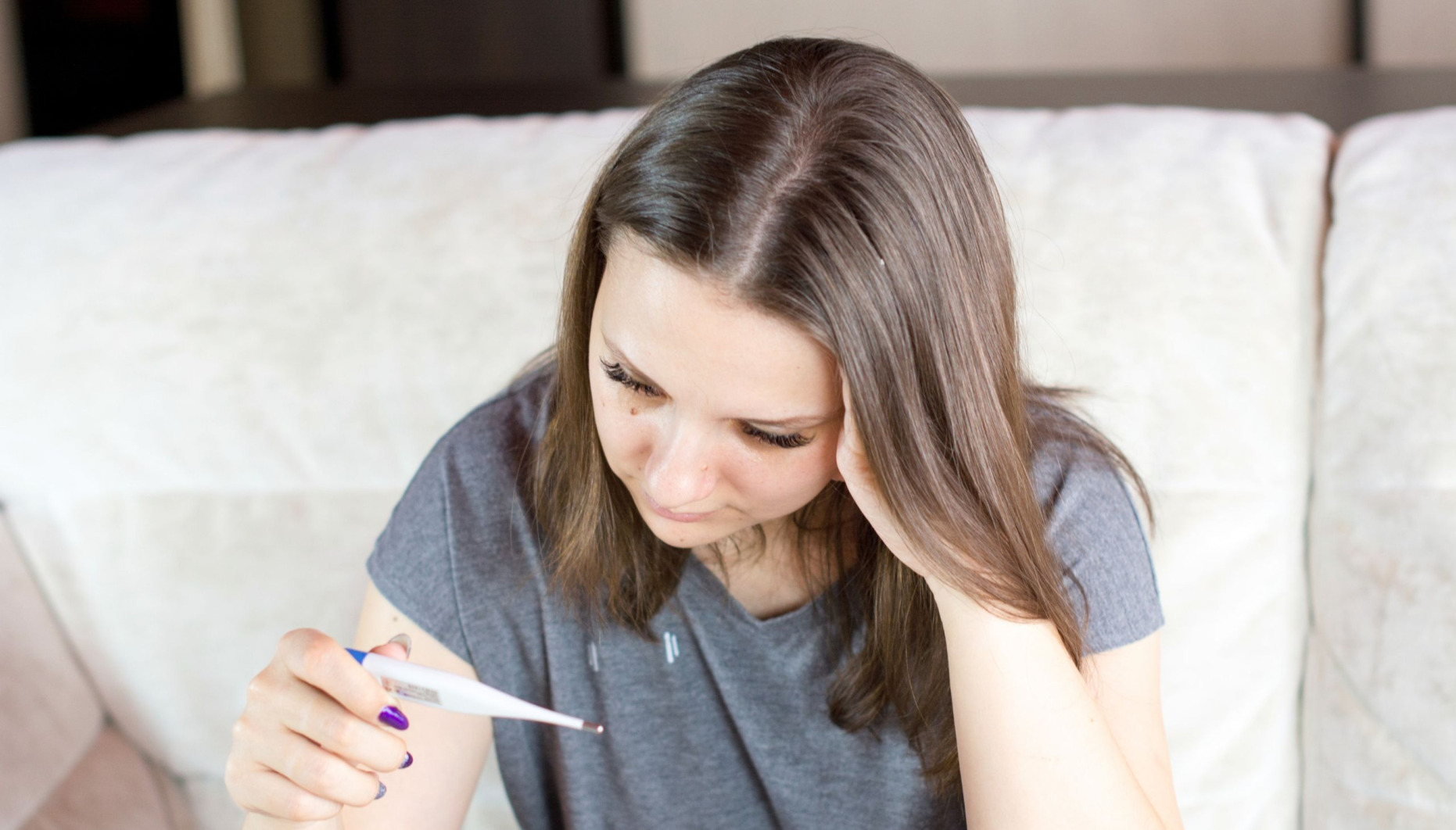 Woman sittting on a sofa holding an electronic thermometer which she's reading.