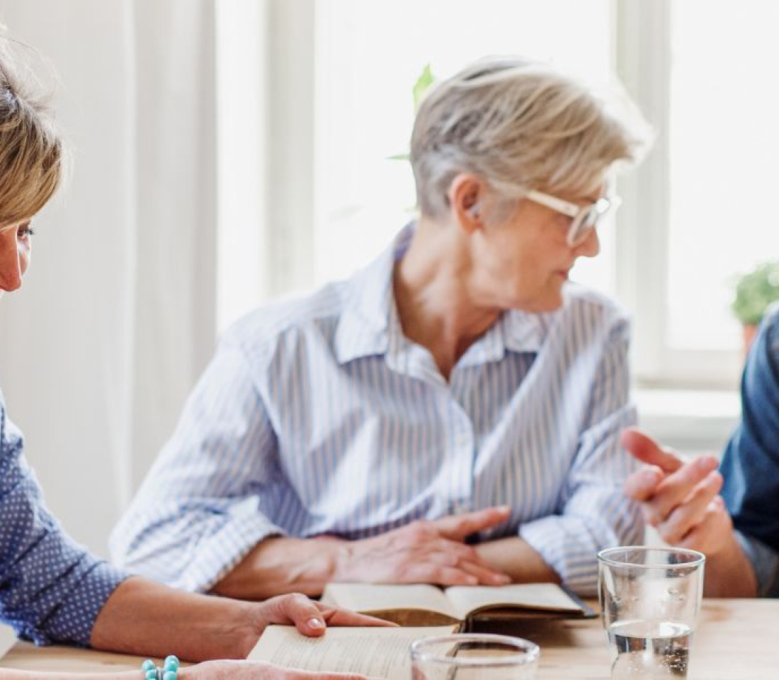 A group of older people sitting around a table with open books in front of them. They are engaged in conversation