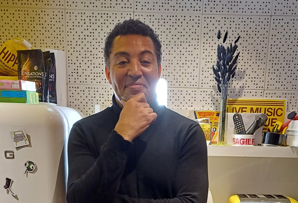 Middle aged man standing in a studio kitchen, looking at the camera and stroking his chin.