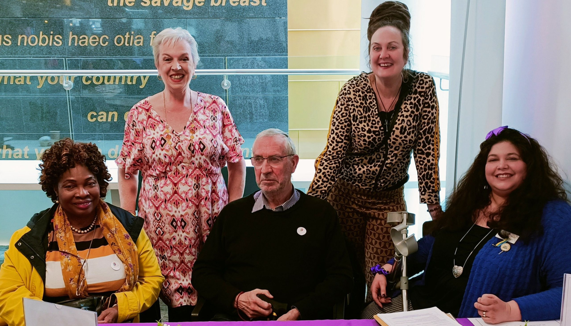 Three people sitting at a table, there are two women standing behind and they are all posing for a photograph