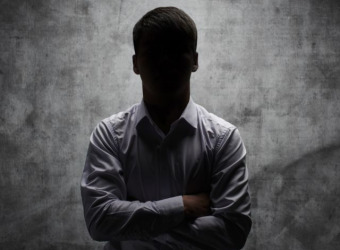 A dimly lit silhouette of a man standing against a grey background with his arms folder