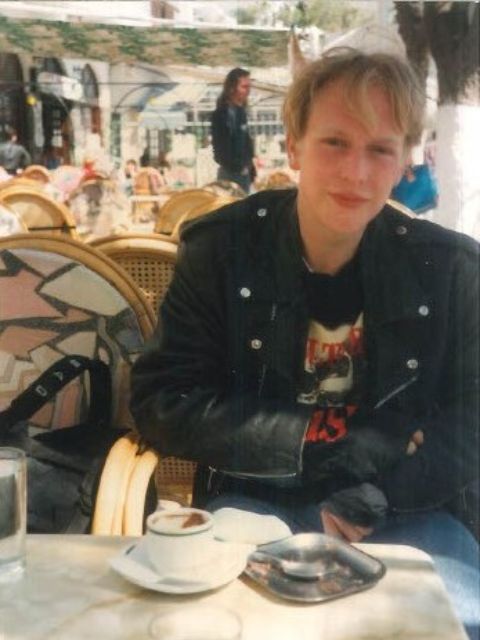 Gareth when he was a younger man.  He is wearing a black leather jacket and sitting at a café table outside in the sunshine.