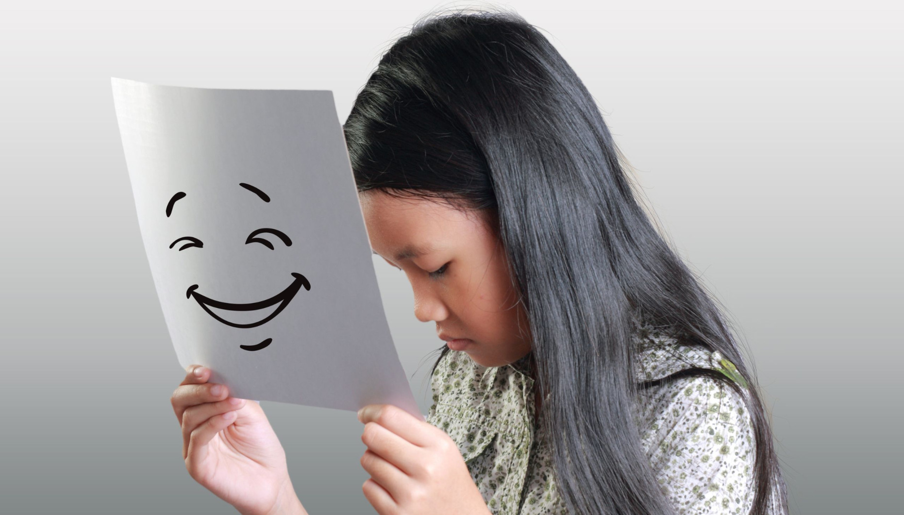 A sad young girl holding a piece of paper in front of her face. There is a smiling face drawrn on the paper to illustrate that she is masking her real feelings