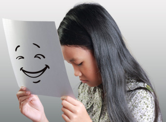 A sad young girl holding a piece of paper in front of her face. There is a smiling face drawrn on the paper to illustrate that she is masking her real feelings