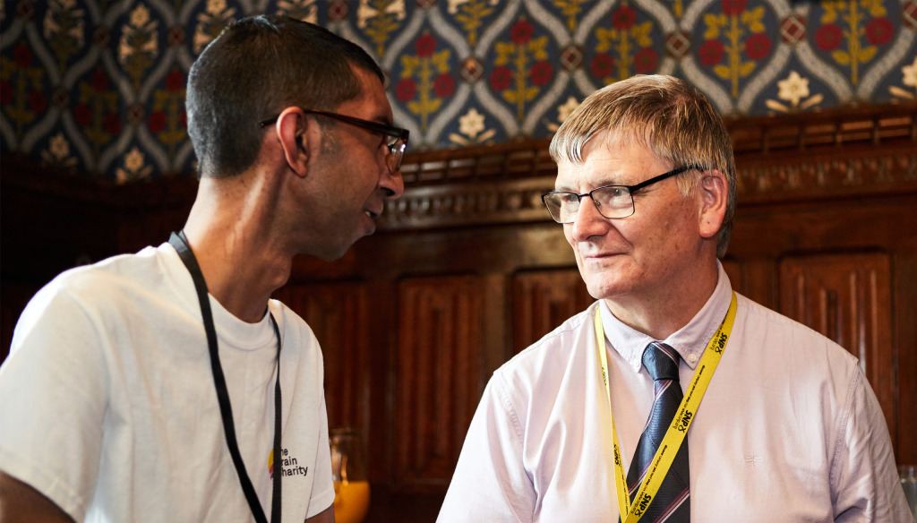 Two men speaking in a wood-panelled room. ONe is wearing a Brain CAHrity T-shirt & one  a shirt and tie