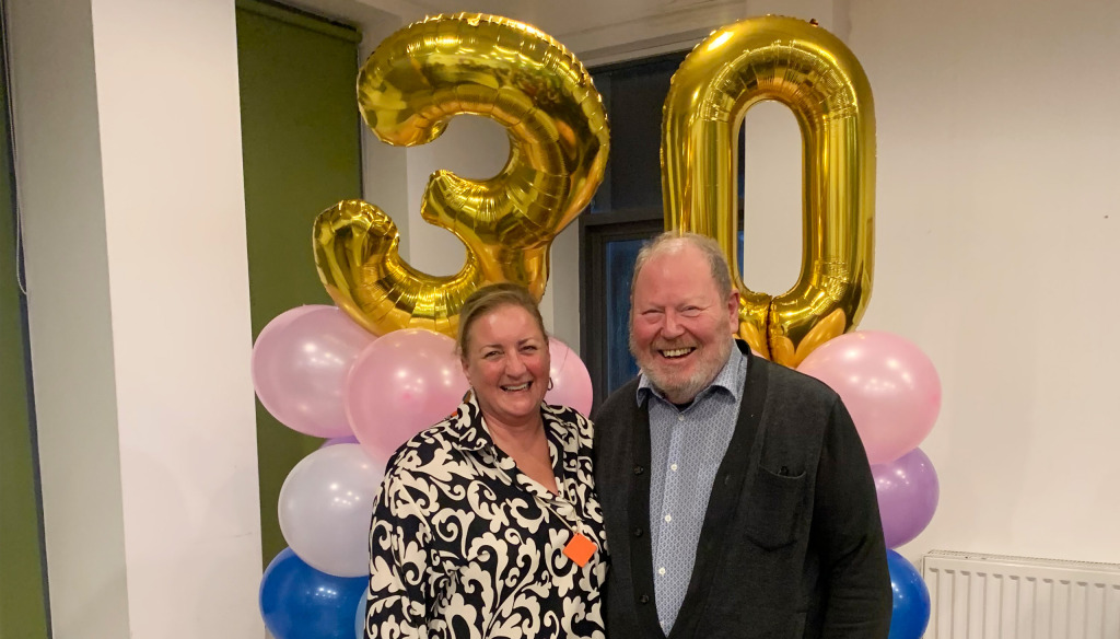 A woman and a man stanging in front of a baloon arch with the number thirty in baloons. They are smiling at the camera 