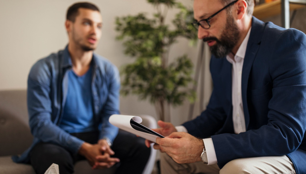 An older man listening to a younger man speaking. They are both seated. The older man is looking at his notes