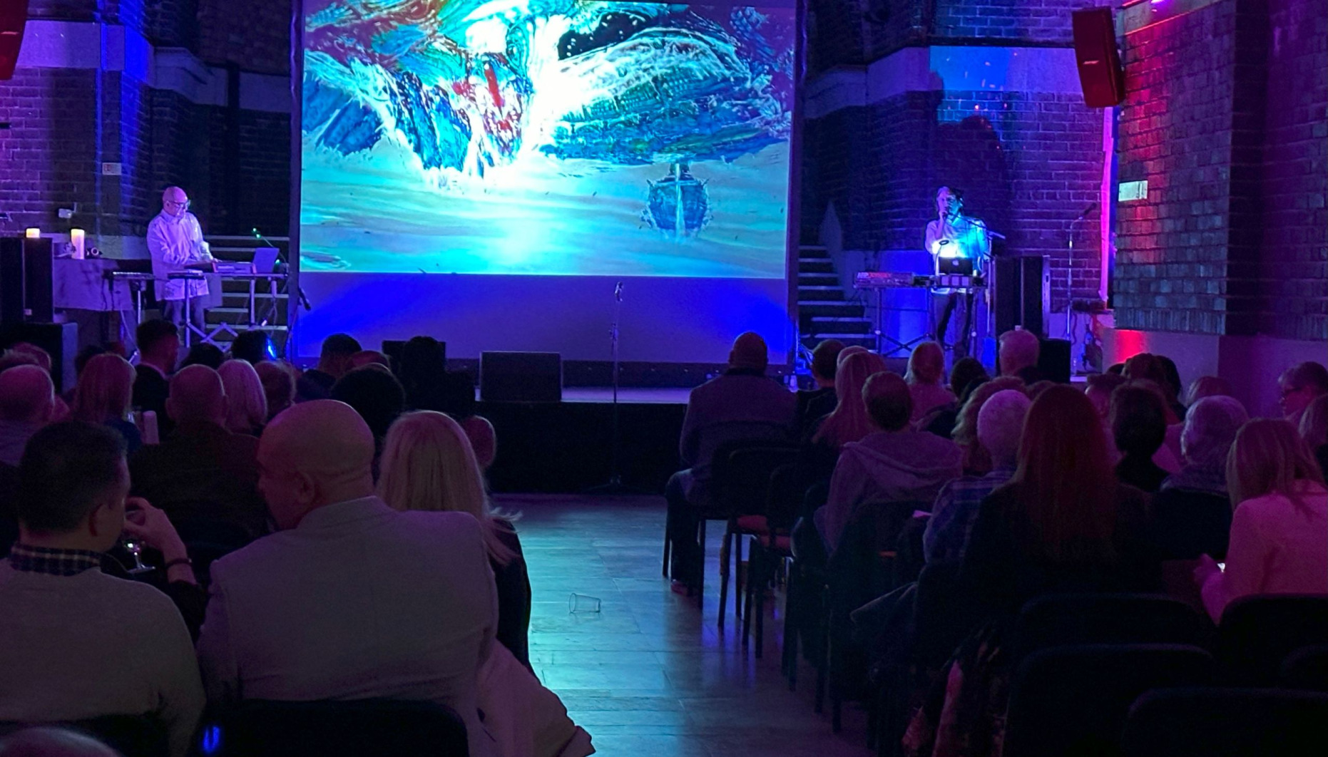 2 men with synthesisers perfoem on a stage with slideshow in background