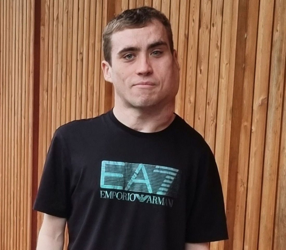 A young man in black T-shirt stands in front of an exterior wall made from wooden cladding.