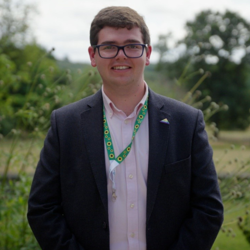 A young man in a shirt and jacket. He is wearing glasses and has a sunflower hidden disability lanyard around his neck. He is smilnig at the camera.