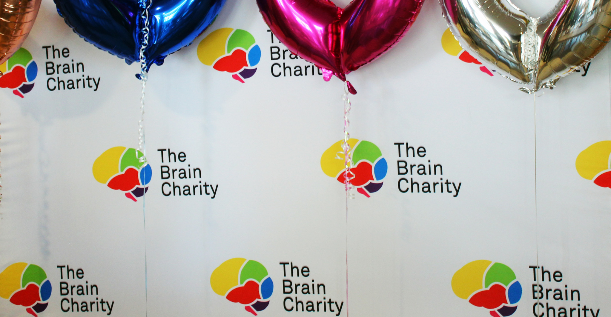 A cloth backdrop with The Brain Charity logo repeated on it. The bottom of some colourful balloons is at the top and ribbons hang down.