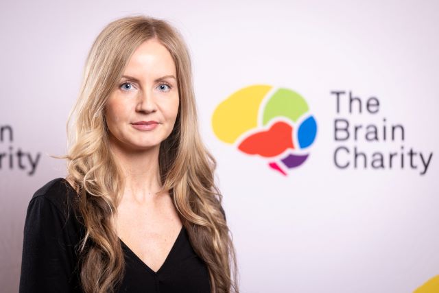 Headshot of Stephanie Hollywood smiling at the camera against a background with Brain Charity logos