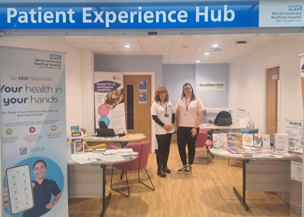 Two women standing in a room with tables, chairs and information leaflets. The sign above the door reads: Patient experience Hub