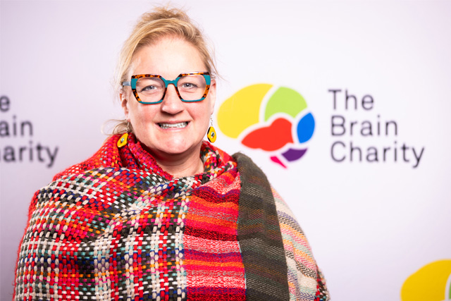 Headshot of Pippa Sargent wearing colourful glasses and shawl. She is smiling at the camera against a background with Brain Charity logos