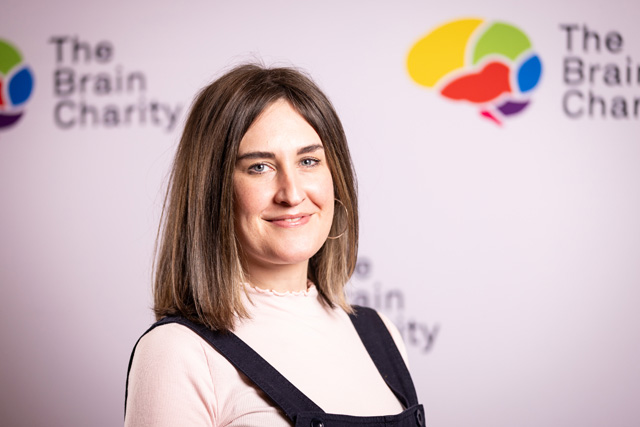 Headshot of Amy smiling at the camera against a background with Brain Charity logos
