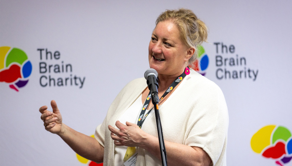 Pippa, a middle aged blond woman in a white top, talking in front of a Brain Charity logo wall.
