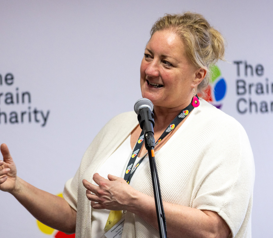 Pippa, a middle aged blond woman in a white top, talking in front of a Brain Charity logo wall.