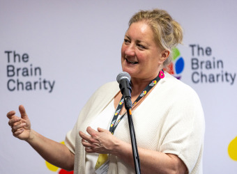 Pippa, a middle aged blond woman in a white top, talking in front of a Brain Charity logo wall.