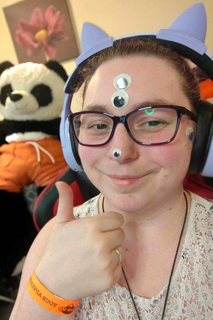 A smiling gamer girl wearing cat ear headphones and giving a thumbs up. She also has three stick on eyes on her face.