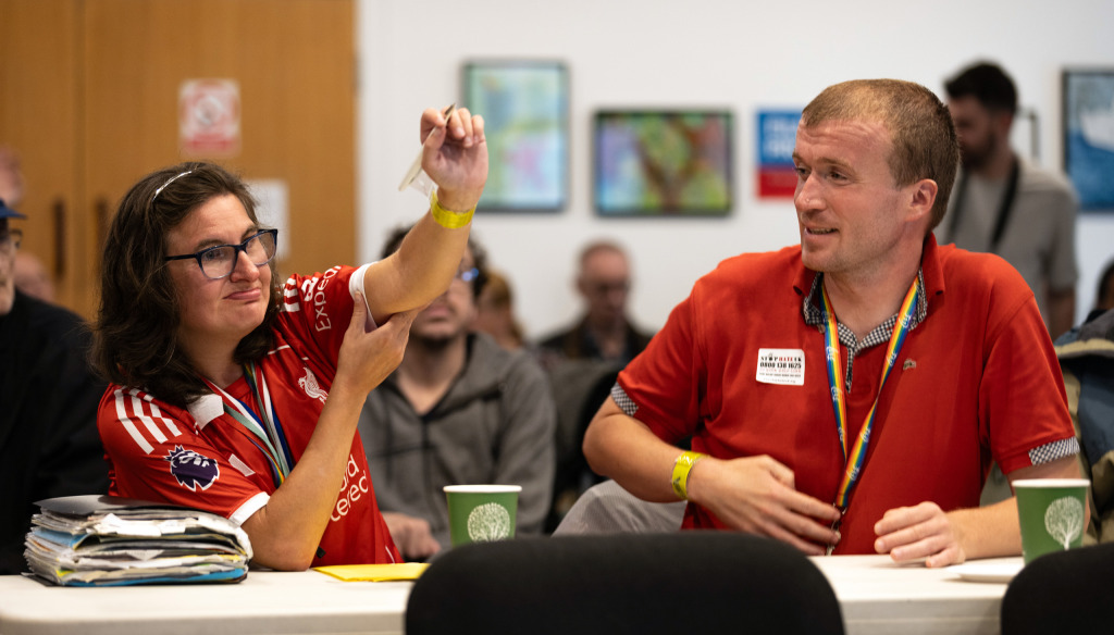 A young woman wearing a red football shirt raises her hand. She is sitting with others in a meeting room at The Brain Charity. A young man is seated beside her looking on.