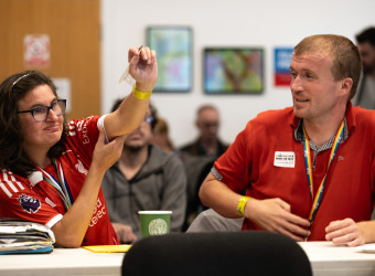 A young woman wearing a red football shirt raises her hand. She is sitting with others in a meeting room at The Brain Charity. A young man is seated beside her looking on.