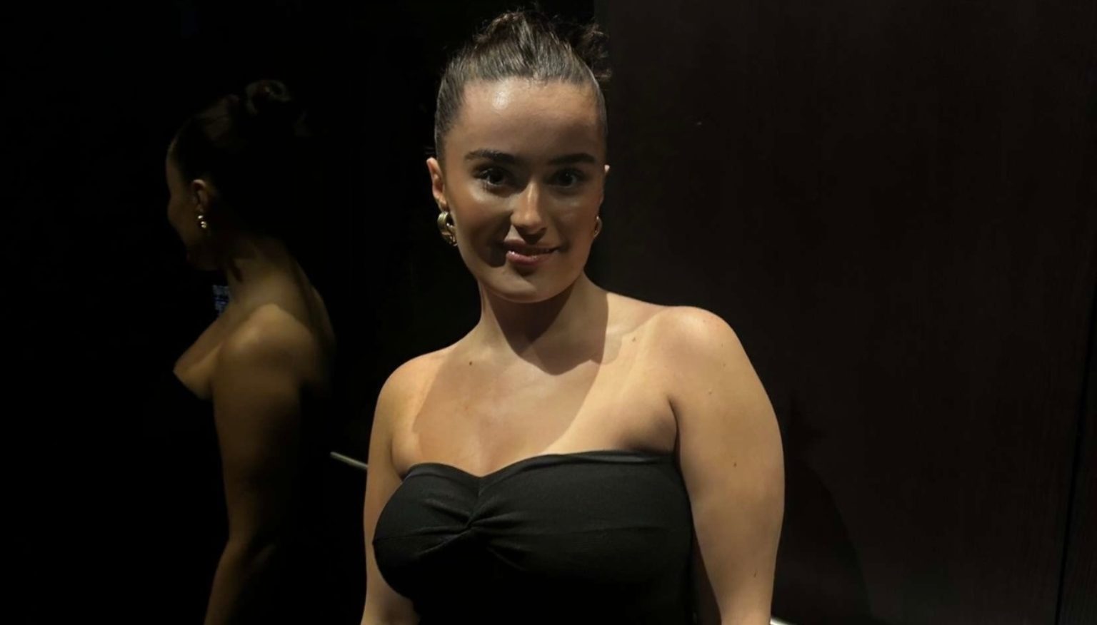A young woman, Aisha, wearing an off the shoulder black dress and looking at the camera