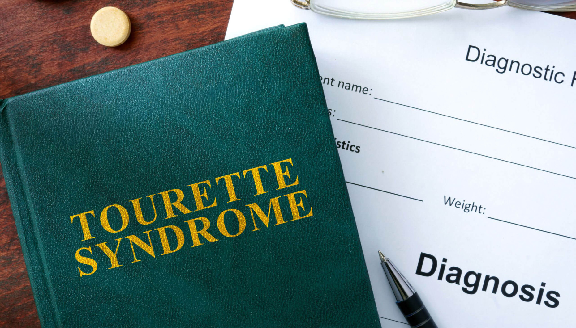A book on a desk with a diagnosis form, pen and glasses. The title of the book is Tourette syndrome.