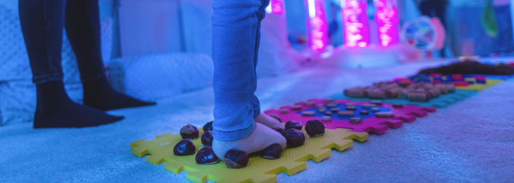 Child walking across foam pads in stockinged feet with different items on. They are currently standing on conkers.