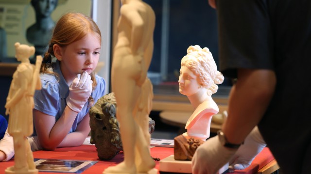 A young person examining classical-style sculptures during a hands-on museum workshop, with small statue replicas and artefacts arranged on a table and an adult facilitator nearby.
