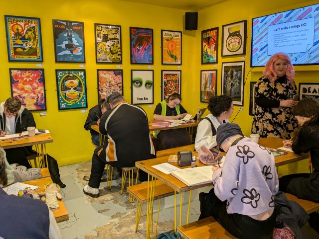 Young people seated at long tables taking part in a comics drawing workshop in a bright yellow room, with illustrated posters on the walls and a presenter standing beside a screen showing a slide at the front