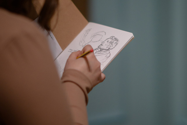 A person sketching with a pencil in a sketch book. We are looking over their shoulder.
