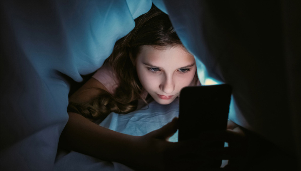 A girl hiding under the covers and using her mobile. The light from the screen illuminates her face.