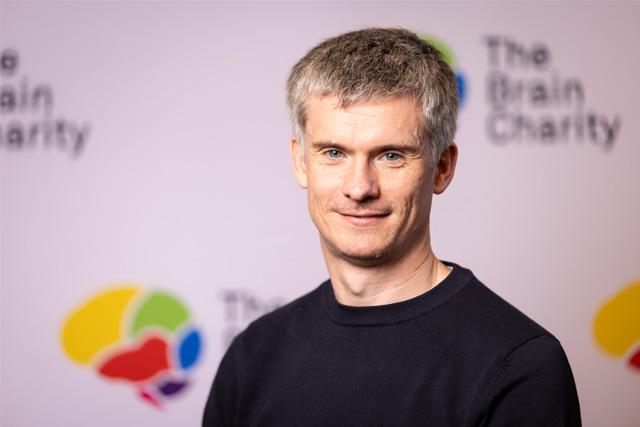 Headshot of Matt Meaney smiling at the camera against a background with Brain Charity logos