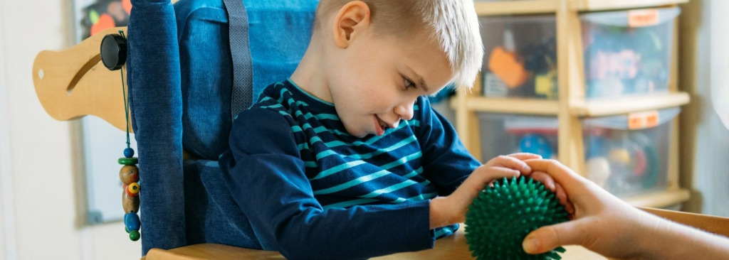 A young boy sitting in a chair and touching a spikey rubber ball held by a man