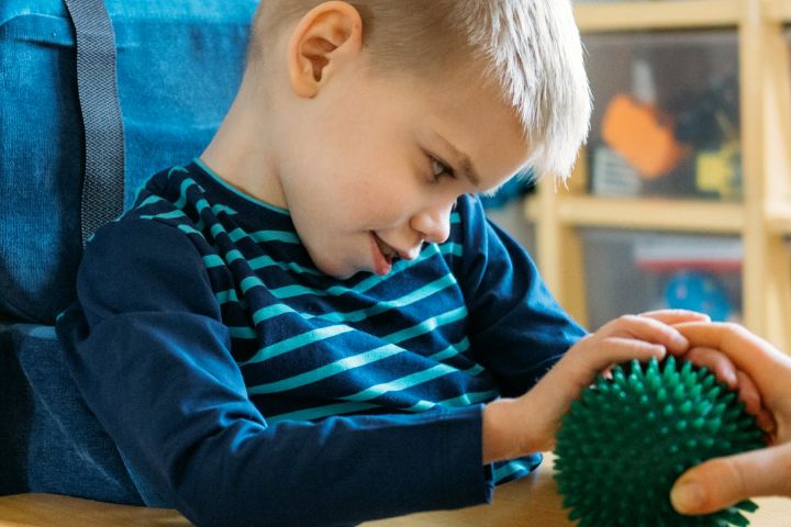 A young boy sitting in a chair and touching a spikey rubber ball held by a man