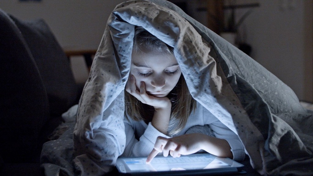 Child reading tablet under covers in bed at night