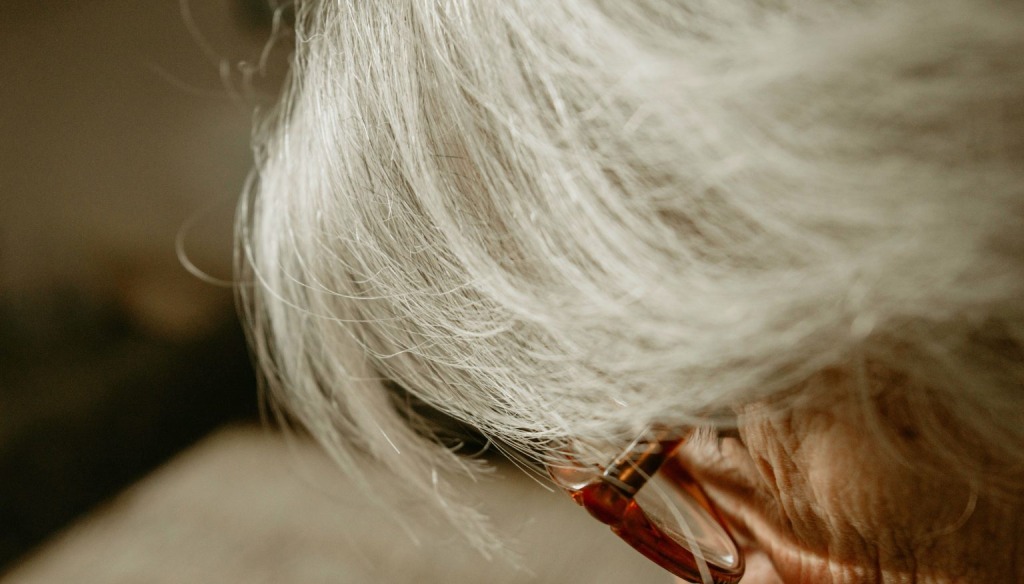 An older person with grey hair and glasses looking down in a close-up portrait focussing on the side of the face.